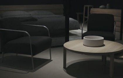 Living room setting with two black armchairs and a round light wood coffee table, with a bedroom in the background.