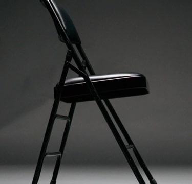 Black folding chair with simple padding, displayed in a dark setting.
